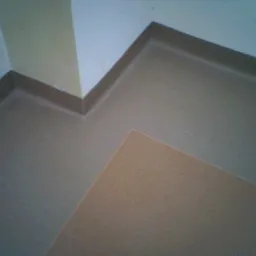 Szczegółowe ujęcie narożnika pomieszczenia z nową, jednolitą posadzką z PVC w odcieniach beżu i brązu, z widoczną listwą przypodłogową w kolorze ciemnego brązu.